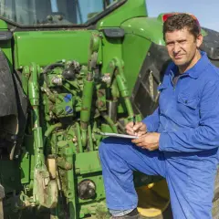 Bauer in blauer Latzhose prüft Unterlagen neben seinem Traktor. Er plant die nächste Ernte und denkt über den **Einkommensteuer Landwirtschaft** nach.