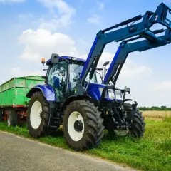Blauer Traktor mit Frontlader und grünem Anhänger steht am Feldrand. Ein Traktor fahren ohne Landwirtschaft ist hier möglich, vielleicht für eine Baustelle.
