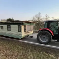 Ein roter Traktor zieht einen Wohnwagen über eine kurvige Straße. Sie sind mit Traktor und Wohnwagen unterwegs, um ein neues Zuhause zu finden.