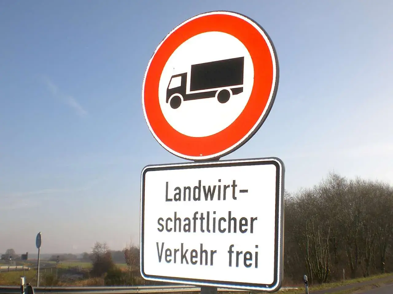 Verkehrsschild: LKW verboten, aber landwirtschaftlicher Verkehr frei. Blaue Himmel und Bäume im Hintergrund.