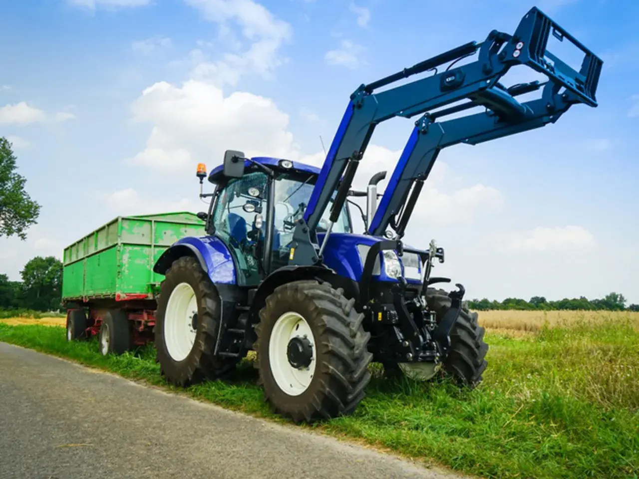 Blauer Traktor mit Frontlader und grünem Anhänger steht am Feldrand. Ein Traktor fahren ohne Landwirtschaft ist hier möglich, vielleicht für eine Baustelle.
