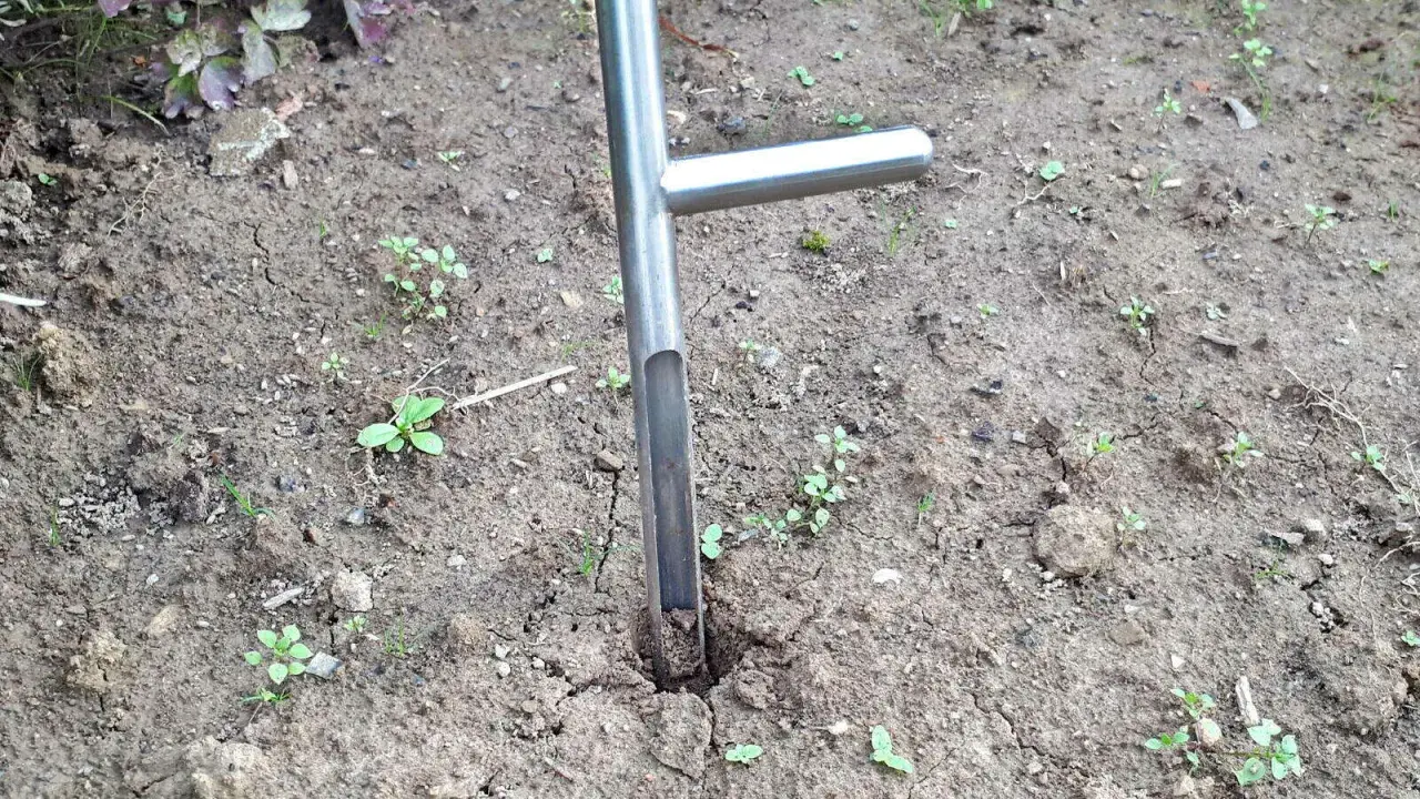 Ein Werkzeug zum Bodenproben ziehen steckt im trockenen Erdreich. Kleine grüne Pflänzchen sprießen zwischen den Rissen im Boden.