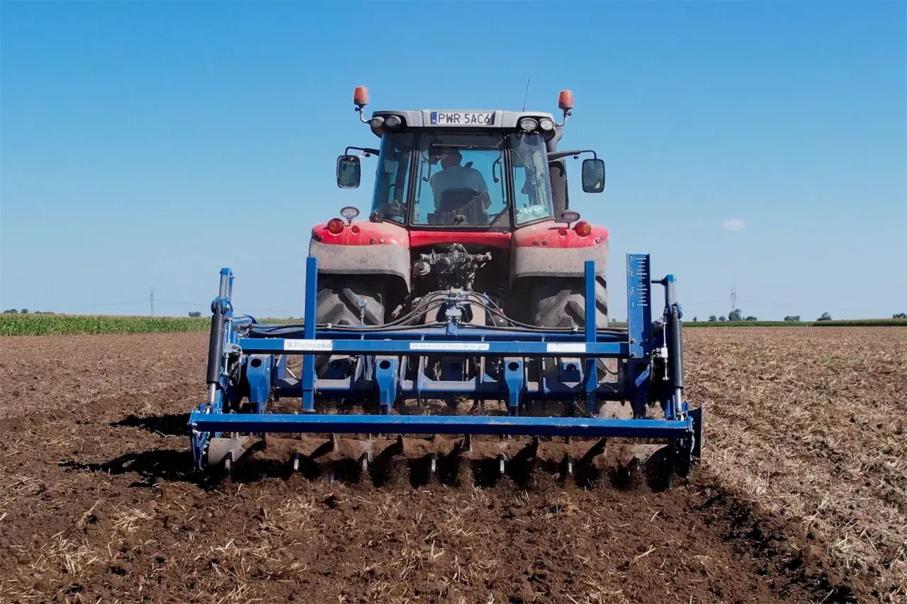Traktor mit einem blauen Gerät für pfluglose Bodenbearbeitung auf einem Feld unter blauem Himmel.