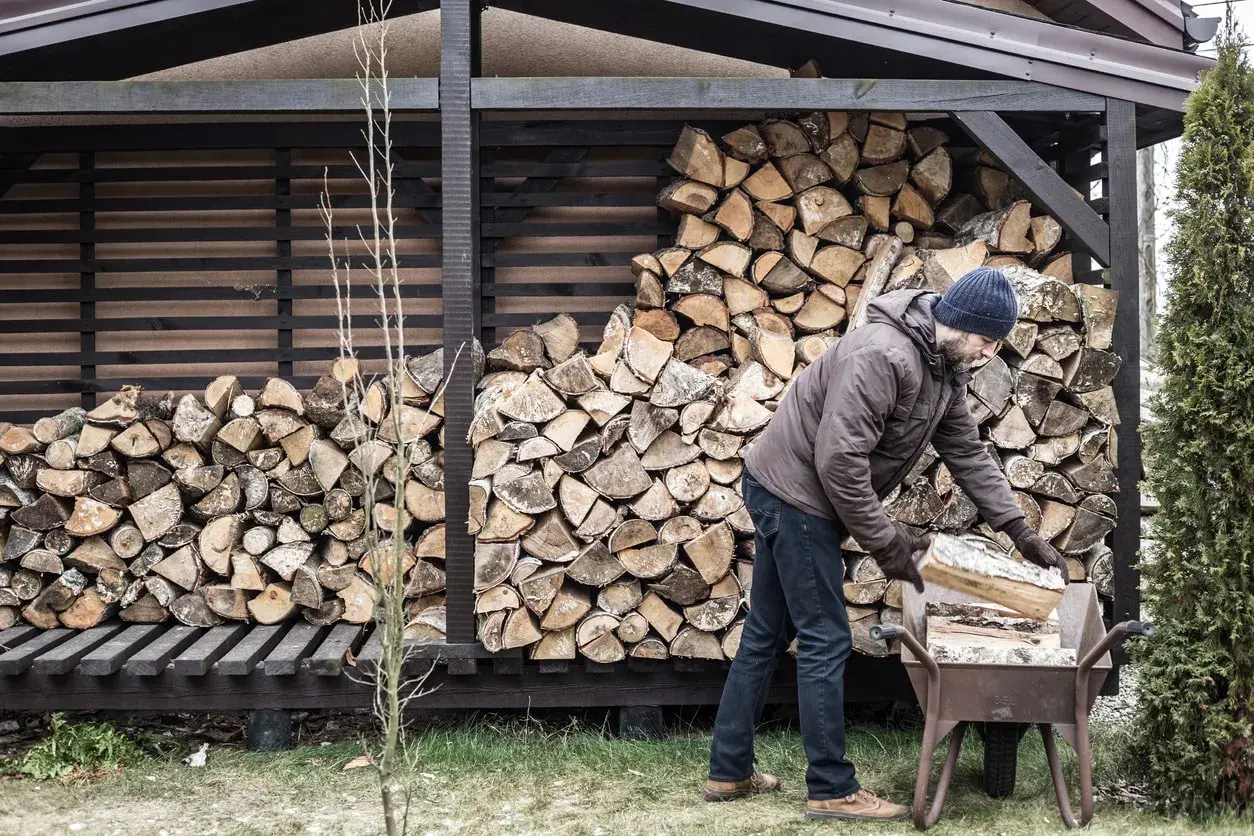 Mann mit blauer Mütze lagert Holz in einer Schubkarre vor einem Holzschuppen.