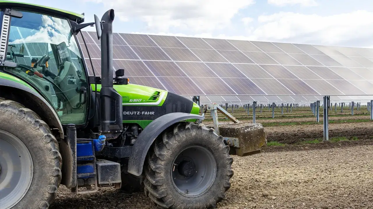 Grüner Traktor vor einer Solaranlage. Die agri PV voraussetzungen werden hier sichtbar: Landwirtschaft und erneuerbare Energien vereinen sich.