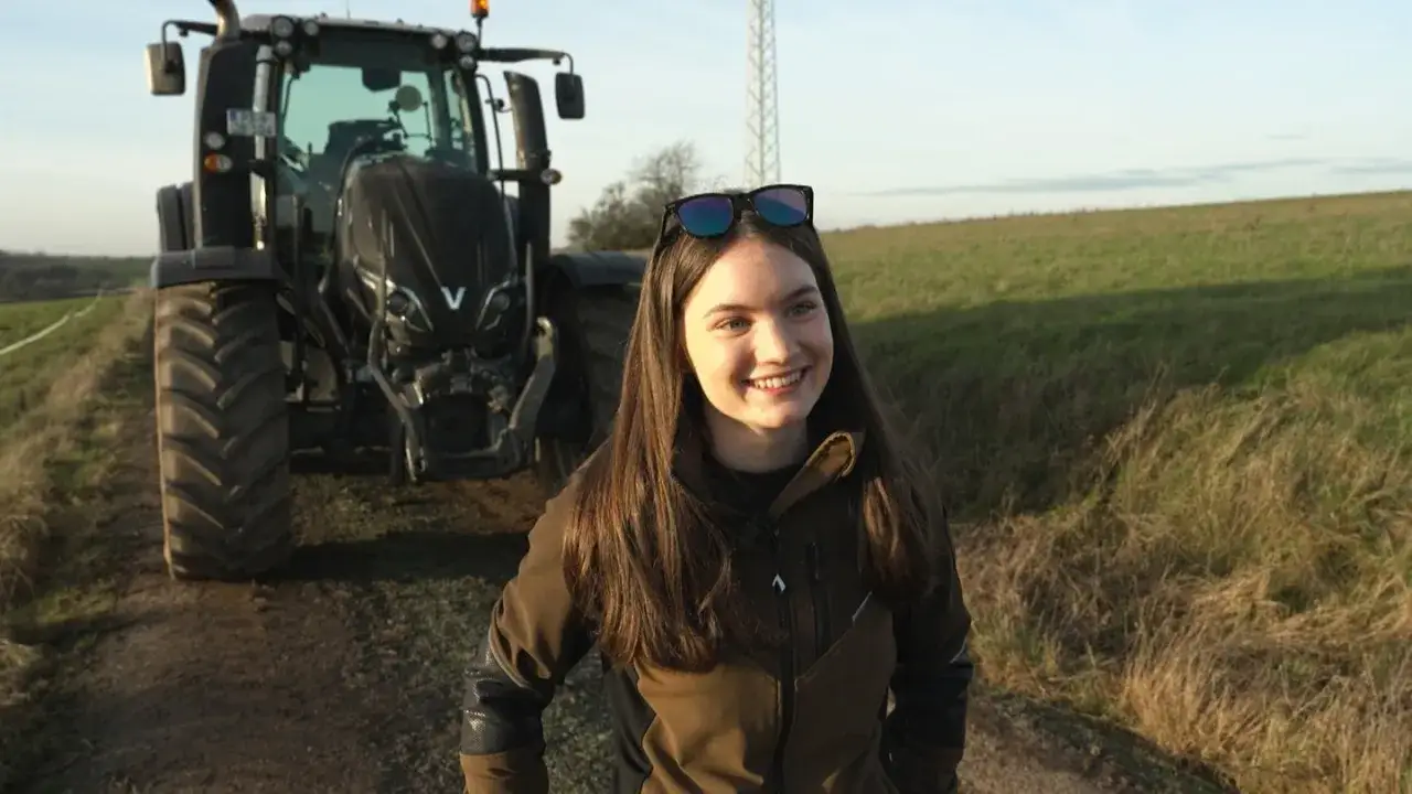 Junge Frau lächelt vor einem großen Traktor. Sie fragt sich, welche Traktoren darf ich mit Klasse B fahren, während sie auf dem Feld steht.