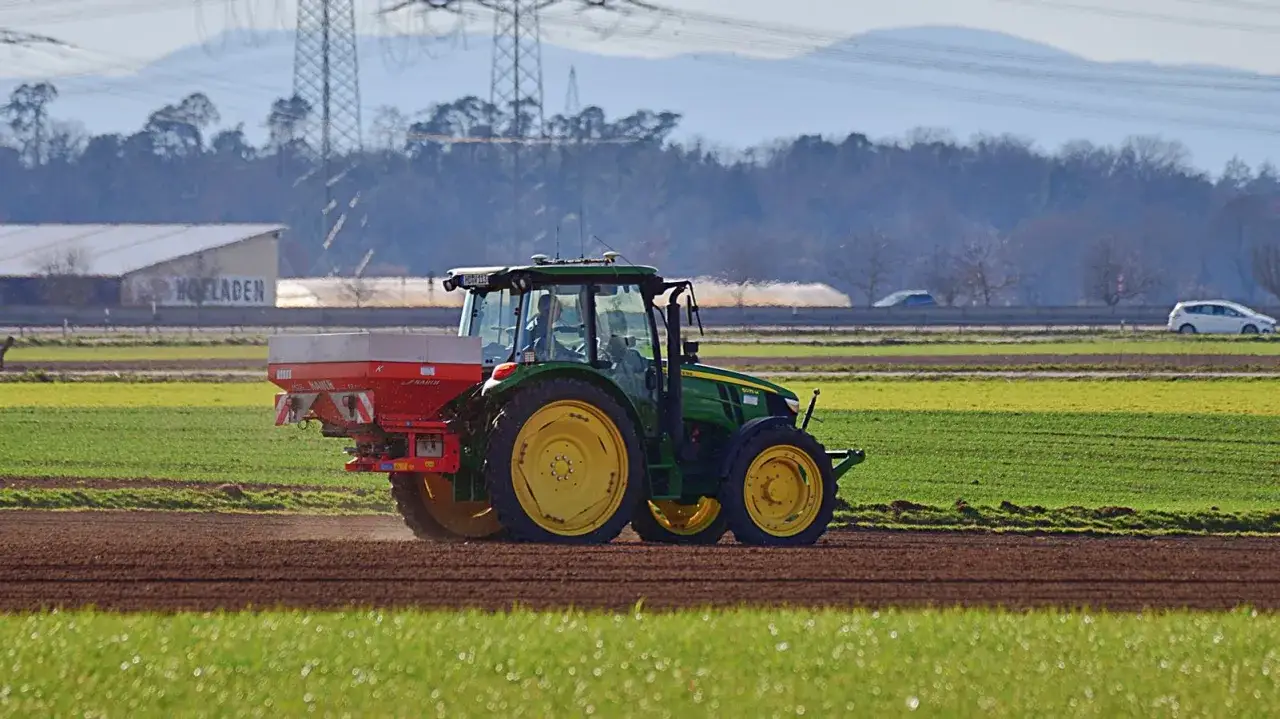 Grüner Traktor mit Düngerstreuer auf einem Feld. Die Entwicklung des Dieselpreises beeinflusst die Landwirtschaft.