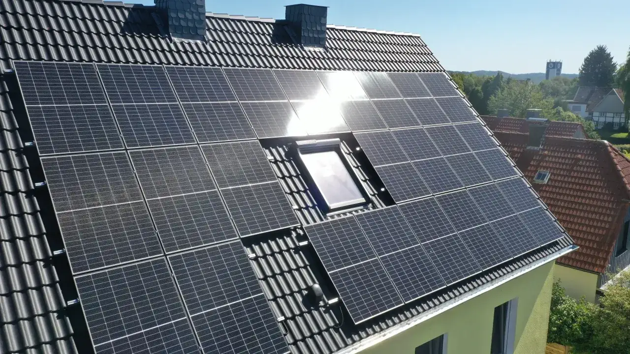 Große Fläche mit solar aufs dach Paneelen auf einem dunklen Ziegeldach, daneben ein Dachfenster und Schornsteine.