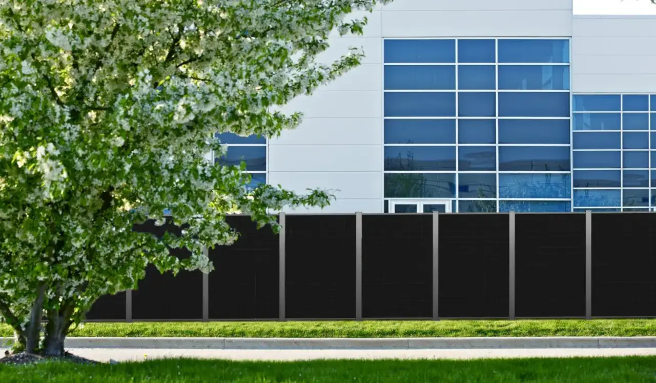 Ein moderner, schwarzer Zaun mit integrierten Solarmodulen vor einem B&uuml;rogeb&auml;ude. Ein bl&uuml;hender Baum im Vordergrund. Ein solarzaun sinnvoll f&uuml;r Energie.