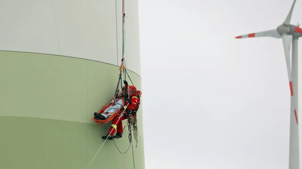 Rettungseinsatz an einem Windrad nach einem Unfall. Ein Retter sichert eine Person in einer Trage, die am Turm herabgelassen wird.