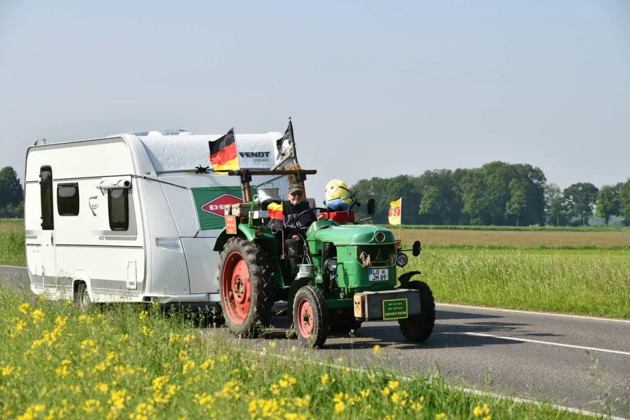 Ein Mann fährt mit einem grünen Deutz-Traktor und einem Wohnwagen auf einer Straße. Ein Minion-Plüschtier sitzt auf dem Traktor.