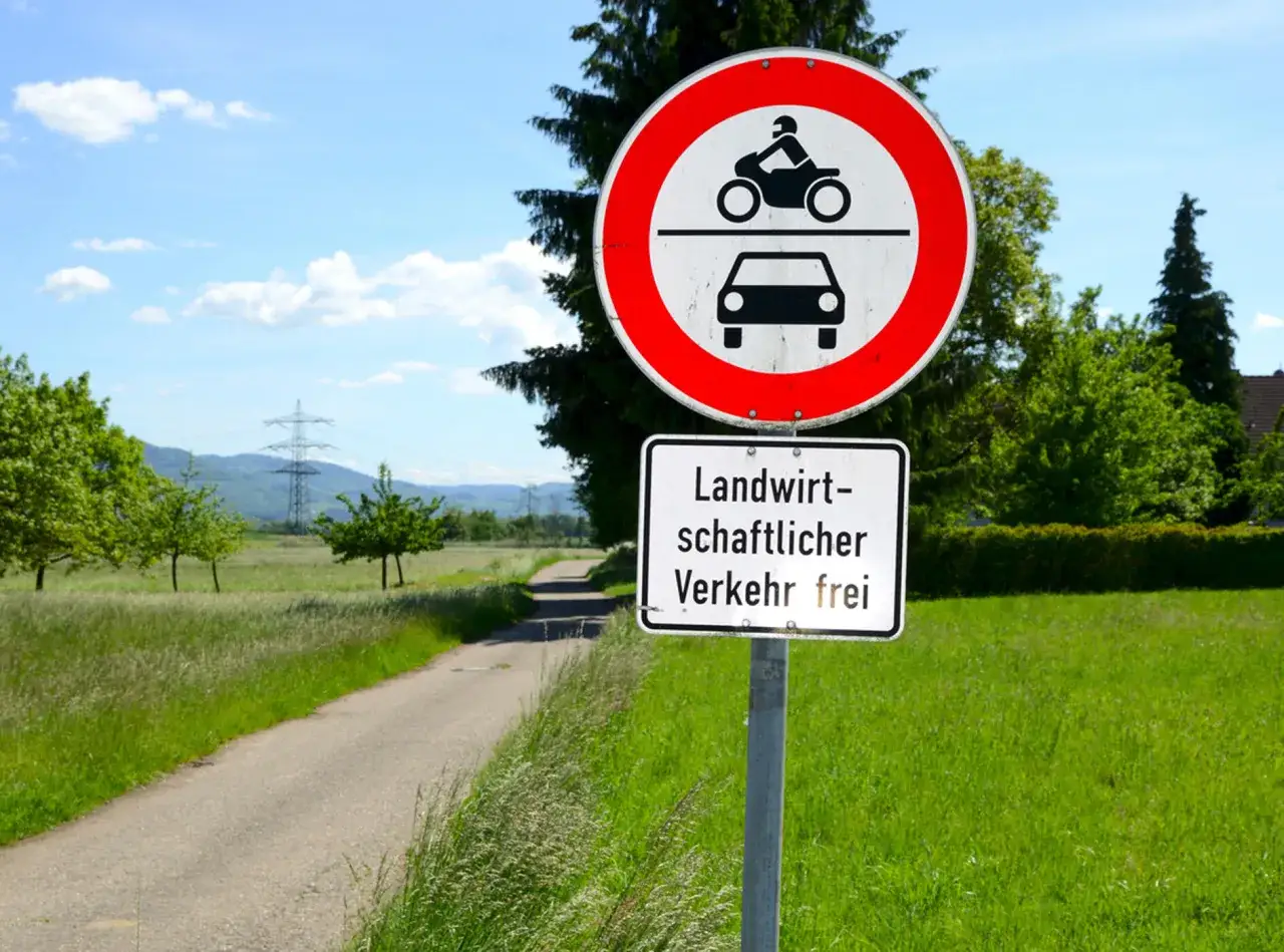 Verkehrsschild: Motorrad und Auto verboten, landwirtschaftlicher Verkehr frei. Feldweg führt durch grüne Wiesen.