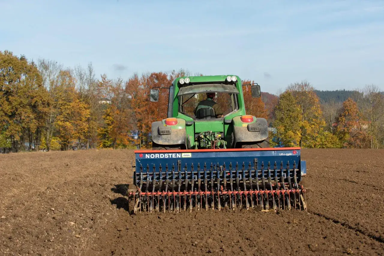 Grüner Traktor mit Sämaschine bereitet Feld für die Sommergerste Aussaat vor. Herbstliche Bäume im Hintergrund.